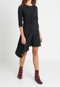 Černé šaty s bílými puntíky, dlouhými rukávy a asymetrickým lemem. Měkká tkanina, mírně zřasená v pase, spárovaná s lesklými červenými kotníkovými botami.