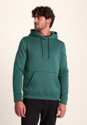 Green hoodie made from a textured material, featuring a front pocket, drawstring hood, and a relaxed fit. Worn with black pants.