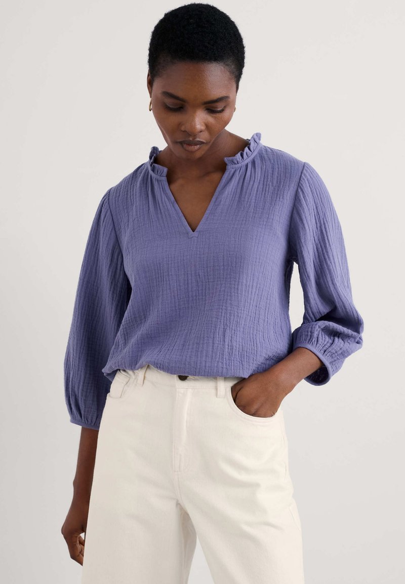 Blusa lavanda con texture, scollo a V e colletto arricciato, dotata di maniche a tre quarti e abbinata a pantaloni bianchi a vita alta.
