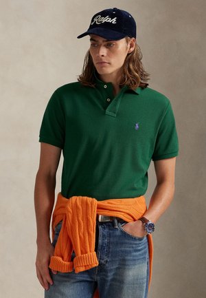 Młody mężczyzna ubrany w ciemnozieloną koszulkę polo, granatową czapkę Ralph, niebieskie dżinsy i pomarańczowy sweter przewiązany wokół talii, z zegarkiem na rękę.