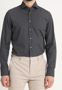 Svart skjorta med knappar och ett litet mönster av vita prickar, långärmad med spetsig krage, ihop med ljusbeige byxor.