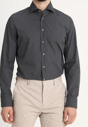 Camicia nera con bottoni, motivo a piccoli pois bianchi, maniche lunghe e colletto a punta, abbinata a pantaloni beige chiaro.