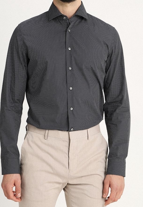 Svart skjorta med knappar och ett litet mönster av vita prickar, långärmad med spetsig krage, ihop med ljusbeige byxor.