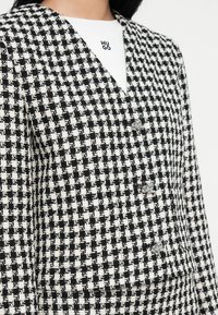 Fekete-fehér houndstooth mintás blézer V-nyakkivágással, dupla gombolású kialakítással és két ezüst színű gombbal az elején.