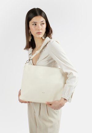 Donna con abito beige tiene una grande borsa spalla di colore avorio con tracolla intrecciata, guarda di lato contro uno sfondo semplice.