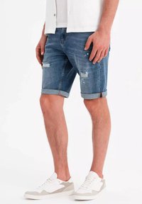 Denimshorts i en ljusblå tvätt, med fransade kanter och slitna detaljer, kombinerade med vita och grå sneakers.