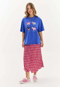 Modrá oversized tričko s grafikou krav a textom, skombinované s červenou sukňou s modrými abstraktnými tvarmi, doplnené o Ružové športové topánky.