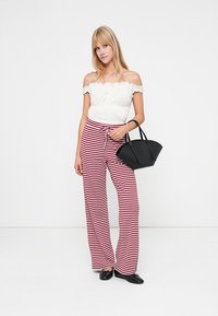 Jovem mulher vestindo uma blusa branca de ombros à mostra e calças listradas vermelhas e brancas, segurando uma bolsa preta, em pé contra um fundo branco liso.