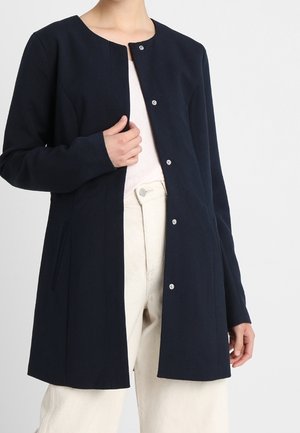 Femme portant un manteau boutonné bleu marine à manches longues par-dessus un haut blanc et un pantalon beige clair, la main tenant l'avant du manteau près de la poitrine.