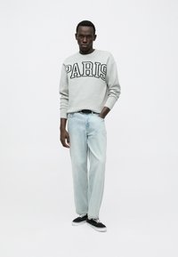 Pull gris avec l'inscription "PARIS" en lettres noires, jean bleu clair, et baskets noires à semelles blanches. Le mannequin pose les mains dans les poches.