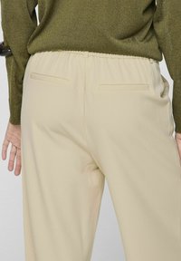 Beige broek met een gladde textuur en een elastische tailleband, voorzien van twee schuine achterzakken en een lichte draping.