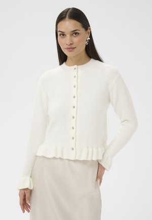 Kobieta z długimi ciemnymi włosami, ubrana w biały kardigan na guziki z falbanką na dole i mankietach, zestawiony z jasnobeżową spódnicą, na jednolitym tle.