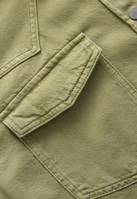 Groene katoenen stof met een gestructureerde afwerking. Beschikt over een patchzak met zichtbare stiksels en een metalen drukknoop.