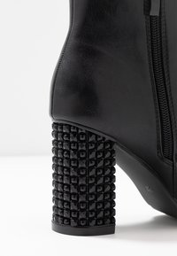 Czarny skórzany but z suwakiem i grubym obcasem pokrytym błyszczącymi kwadratowymi ćwiekami, na jednolitym białym tle.