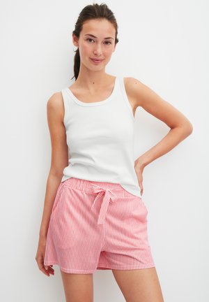 Woman wearing a white sleeveless top and pink and white striped shorts with a tied waist, standing with one hand on her hip against a white background.