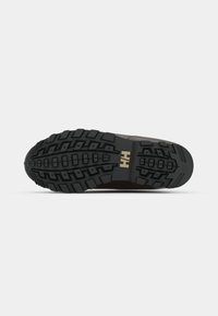Suela de goma negra con surcos profundos, con un logo dorado "HH" y un material superior marrón, texturizado y resistente.