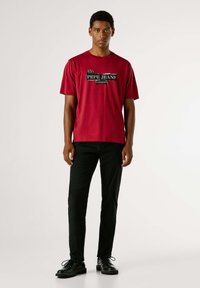 Camiseta roja de algodón de manga corta, con un estampado negro del logo "PEPE JEANS LONDON" en el pecho, combinada con pantalones negros y zapatos.