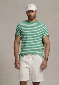 Tricou cu mânecă scurtă, cu dungi verzi și albe, având un mic logo albastru pe partea stângă a pieptului, asortat cu pantaloni scurți bej deschis. O șapcă albă completează ținuta.