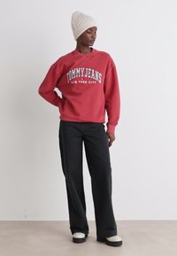 Crvena dukserica s natpisom "TOMMY JEANS NEW YORK CITY", u kombinaciji s crnim širokim hlačama i kremasto-bijelim platformama. Model nosi pletenu kapu.
