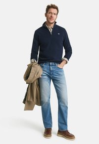 Marinefarbener Pullover mit Viertelreißverschluss und strukturiertem Muster, über einem karierten Hemd getragen, kombiniert mit hellblauen Jeans und braunen Schnürstiefeln.