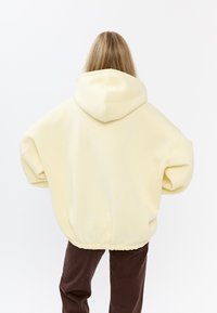 Felpa gialla oversize con orlo regolabile e cappuccio, abbinata a pantaloni marroni. Presenta un tessuto morbido e liscio e maniche esagerate.