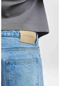 Maglietta grigia infilata in jeans di denim blu chiaro con una patta in pelle beige che mostra la scritta "2Y STUDIOS" sulla vita posteriore.