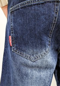 Dark blue denim jeans featuring five-pocket design. Visible branded label on the back pocket, contrasting stitching, and slight fade.