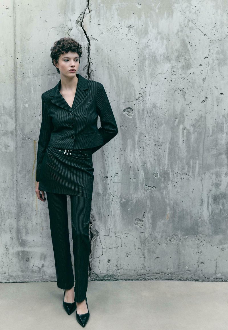Giovane donna con capelli ricci e corti, che indossa un blazer nero a righe sottili, una mini gonna sopra i pantaloni e tacchi neri, in piedi contro un muro di cemento screpolato.