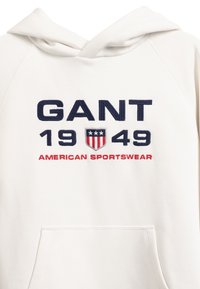 Biała bluza z kapturem z dużym haftowanym logo w granatowym i czerwonym kolorze: "GANT 1949" oraz "AMERICAN SPORTSWEAR" z motywem tarczy. Miękka tkanina.