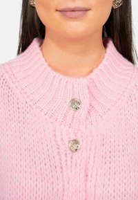 Lichtroze gebreide cardigan met een ribbelkraag en twee decoratieve zilveren knopen. De textuur lijkt zacht en pluizig.