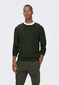 Donkergroene gebreide sweater met een witte kraag eronder. Voorzien van lange mouwen en geribde manchetten. Gecombineerd met olijfgroene cargobroek.