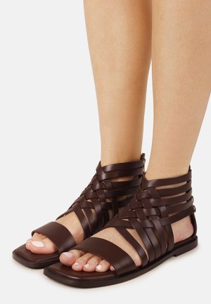 Feet wearing dark brown leather gladiator-style sandals with multiple woven straps and flat square soles against a white background.