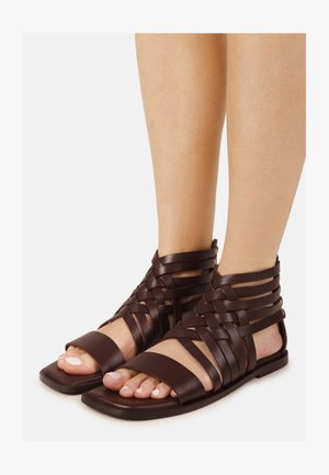 Pies con sandalias de gladiador de cuero marrón oscuro, con múltiples correas tejidas y suelas planas cuadradas, sobre un fondo blanco.