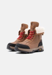 Bruine waterdichte laarzen met namaakbonten boord, rode veters, zwarte rubberen neuskap en Vibram-zool. Stevig ontwerp, geschikt voor buitengebruik.