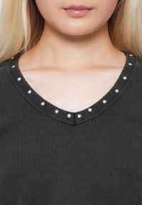 Mørkegrå ribbet topp med V-hals, som har dekorative sølvnagler langs kanten. Stoffet ser mykt ut med en lett tekstur.