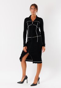 Vestito nero con zip e maniche lunghe, caratterizzato da linee bianche di accento e un orlo spaccato. Texture liscia e design aderente, abbinato a tacchi neri.