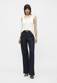 Femme aux cheveux foncés portant un top blanc sans manches, un jean large taille haute foncé, une ceinture noire et des talons ouverts noirs, debout devant un fond blanc.