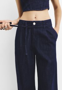 Temno modri jeans hlače z visokim pasom, elastičnim vezanjem, širokimi hlačnicami in stranskimi žepi. Imajo srebrno zaponko na sprednji strani.