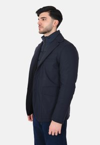 Giacca ibrida blu navy con design strutturato, caratterizzata da un colletto, tasche frontali e una texture liscia, indossata sopra uno strato interno con zip.