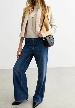 Mujer con suéter beige y chaleco de piel sintética marrón claro, vaqueros azules de pierna ancha, zapatos de tacón negros puntiagudos, collar largo y bolso negro de hombro.