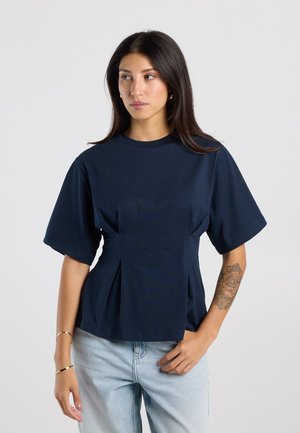 Vrouw met lang donker haar draagt een marineblauw kortärmelig topje en lichtblauwe spijkerbroek, staand tegen een effen grijze achtergrond.