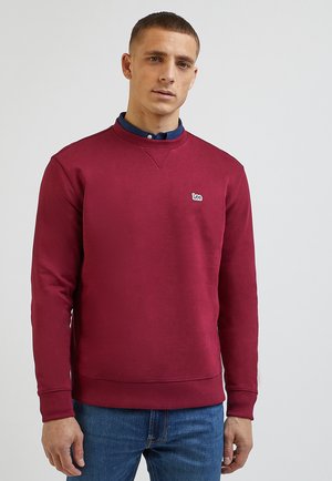 Bordowy sweatshirt z okrągłym dekoltem, z ściągaczami przy rękawach i u dołu, z małym białym logo. Noszony na granatowej koszuli polo, w połączeniu z niebieskimi dżinsami.