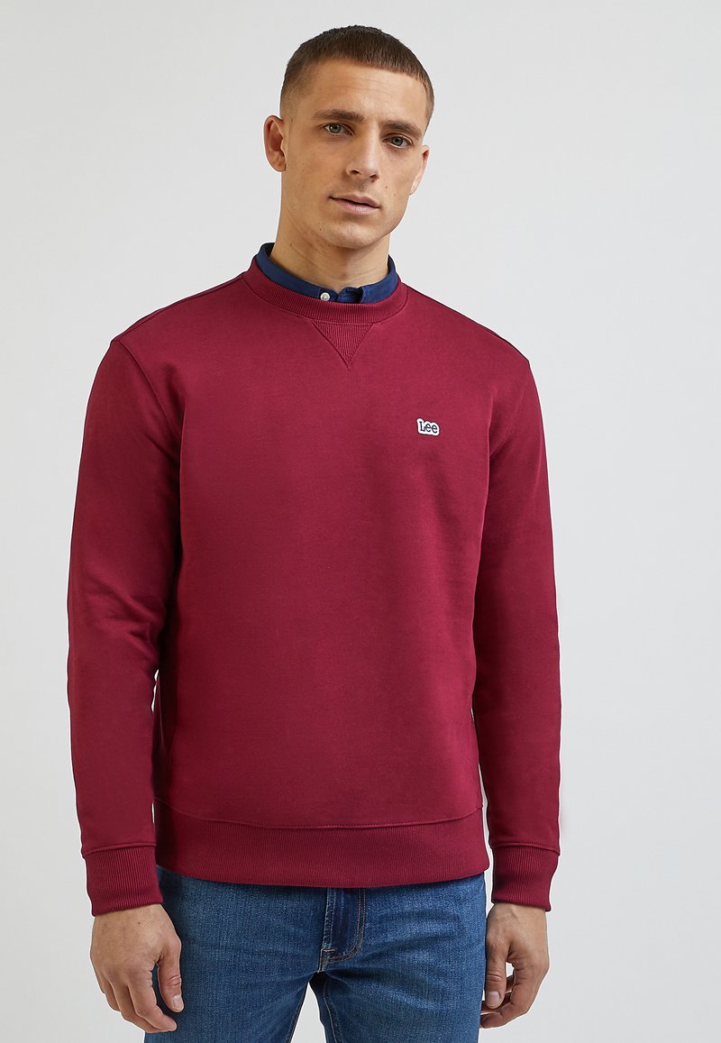 Bordowy sweatshirt z okrągłym dekoltem, z ściągaczami przy rękawach i u dołu, z małym białym logo. Noszony na granatowej koszuli polo, w połączeniu z niebieskimi dżinsami.