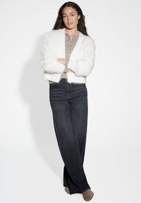 Femme debout les bras croisés, portant une veste blanche en peluche, un haut gris à boutons, un pantalon large bleu marine et des chaussures beiges contre un fond blanc.