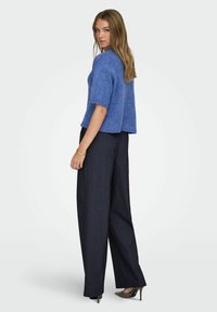 Modrý pletený svetr s krátkými rukávy, kombinovaný s tmavými kalhotami s vysokým pasem a špičatými podpatky. Svetr má volný střih a žebrované okraje.