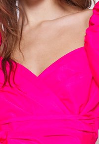 Vrouw in een felroze off-shoulder jurk met een V-halslijn en gerimpeld stofdetail rond de taille.