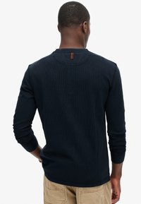 Man wearing navy long-sleeve shirt and beige pants, shown from the back against a plain white background.