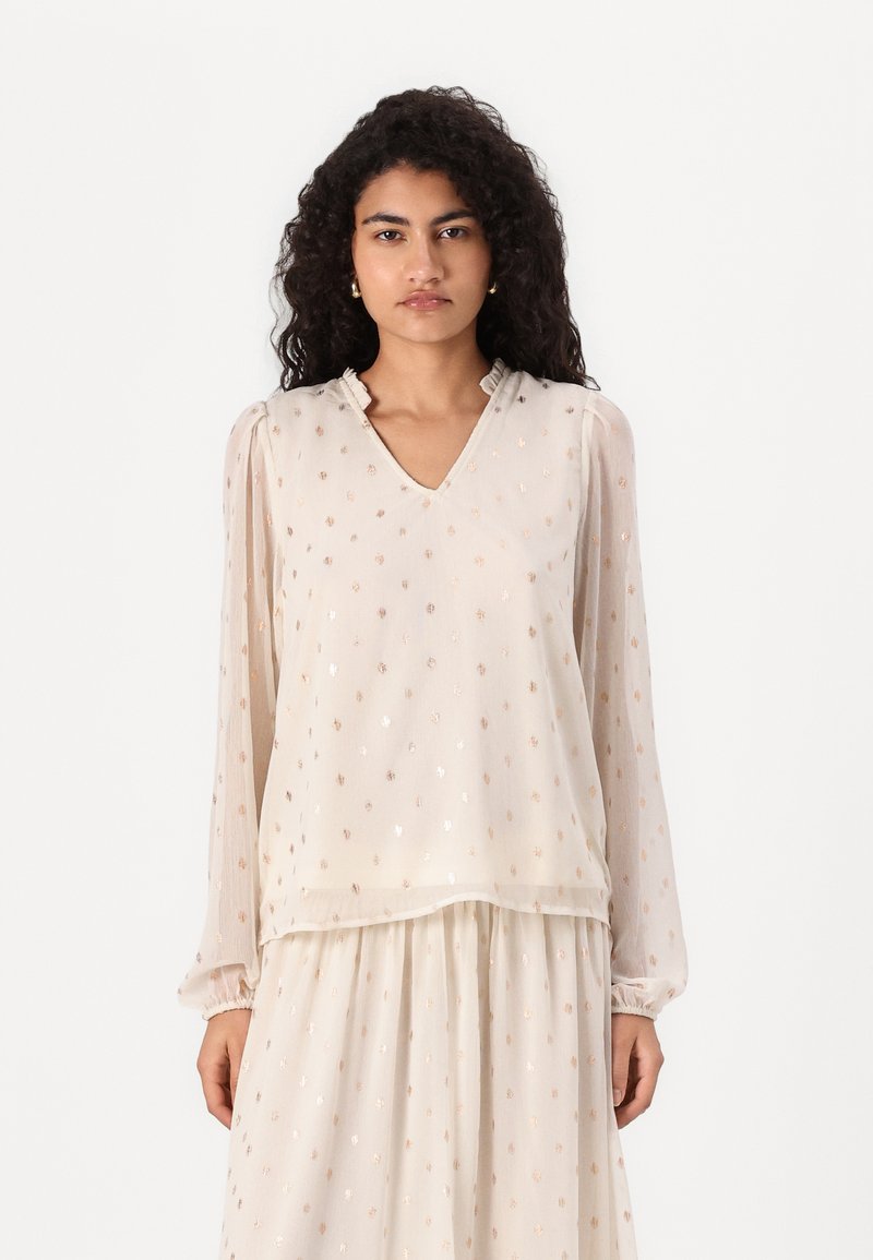 Blusa beige con scollo a V, maniche lunghe e bordi arricciati, caratterizzata da un motivo a pois lucidi su un tessuto trasparente.