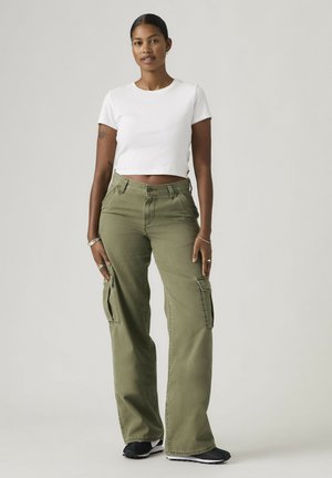 Kobieta stojąca, ubrana w białą krótki top, oliwkowo zielone spodnie typu cargo oraz czarne sneakersy na jednolitym szarym tle.