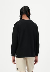 Sweatshirt noir à manches longues avec texture côtelée, col rond et détail de tag sur le côté, associé à un short en velours côtelé beige clair.